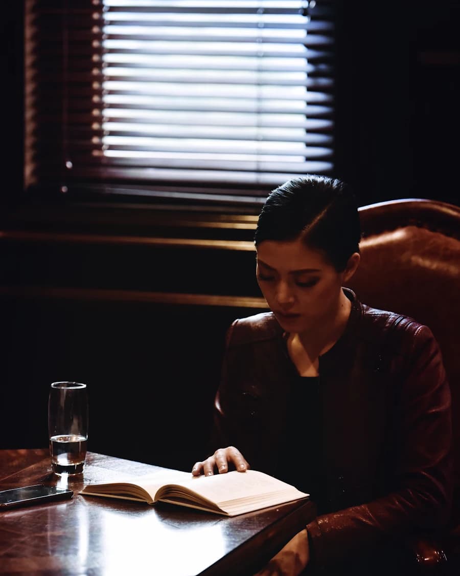 Woman reading a book at a dark wooden desk by window blinds