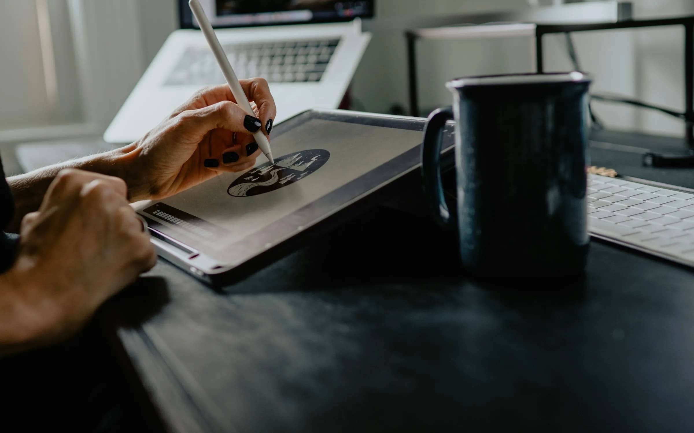 Una mano dibujando en una tableta con un lápiz óptico sobre un escritorio oscuro, con una laptop y una taza al fondo