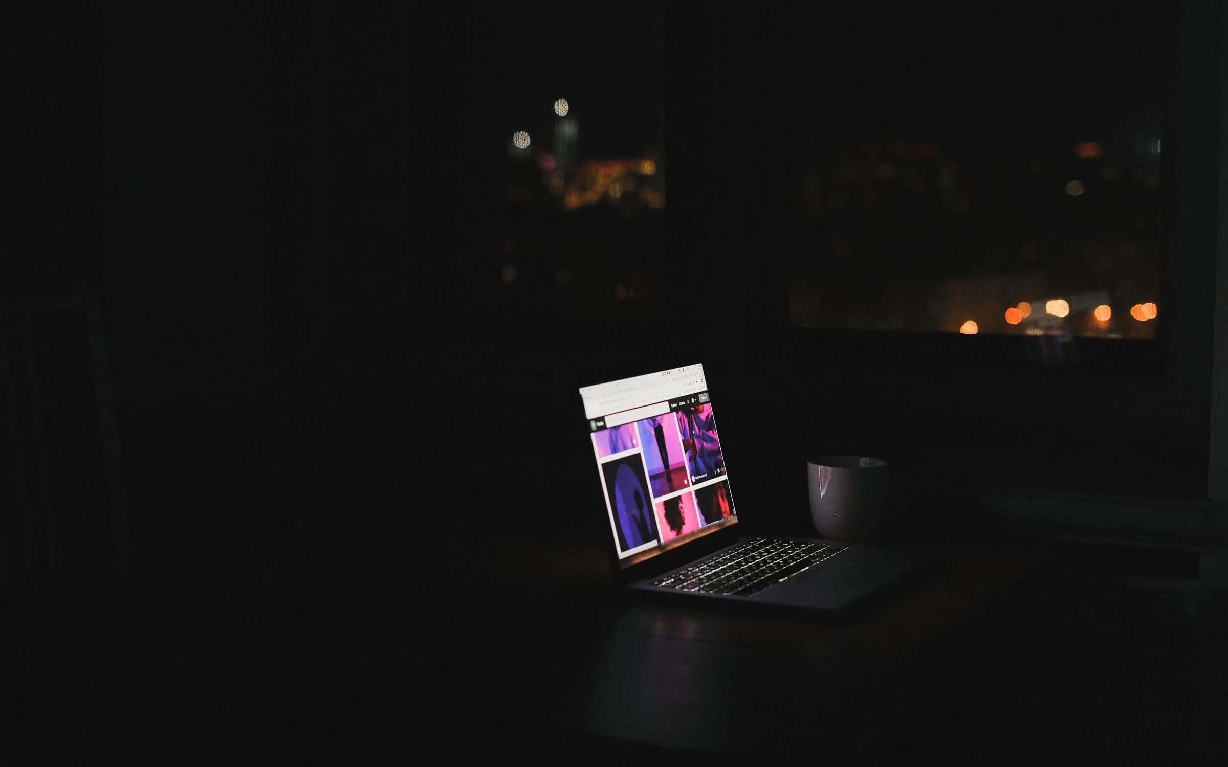 A laptop glowing on a desk in a dark room at night, with blurred city lights through the window