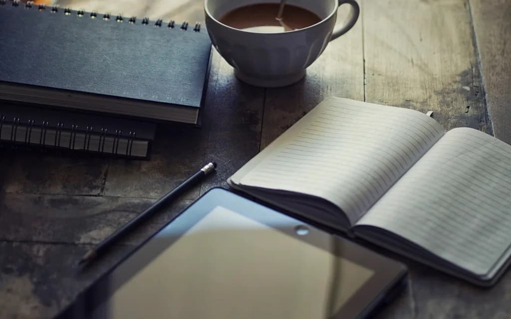 A cup of coffee beside a closed notebook, an open lined journal, a pencil and a tablet on a worn wooden desk