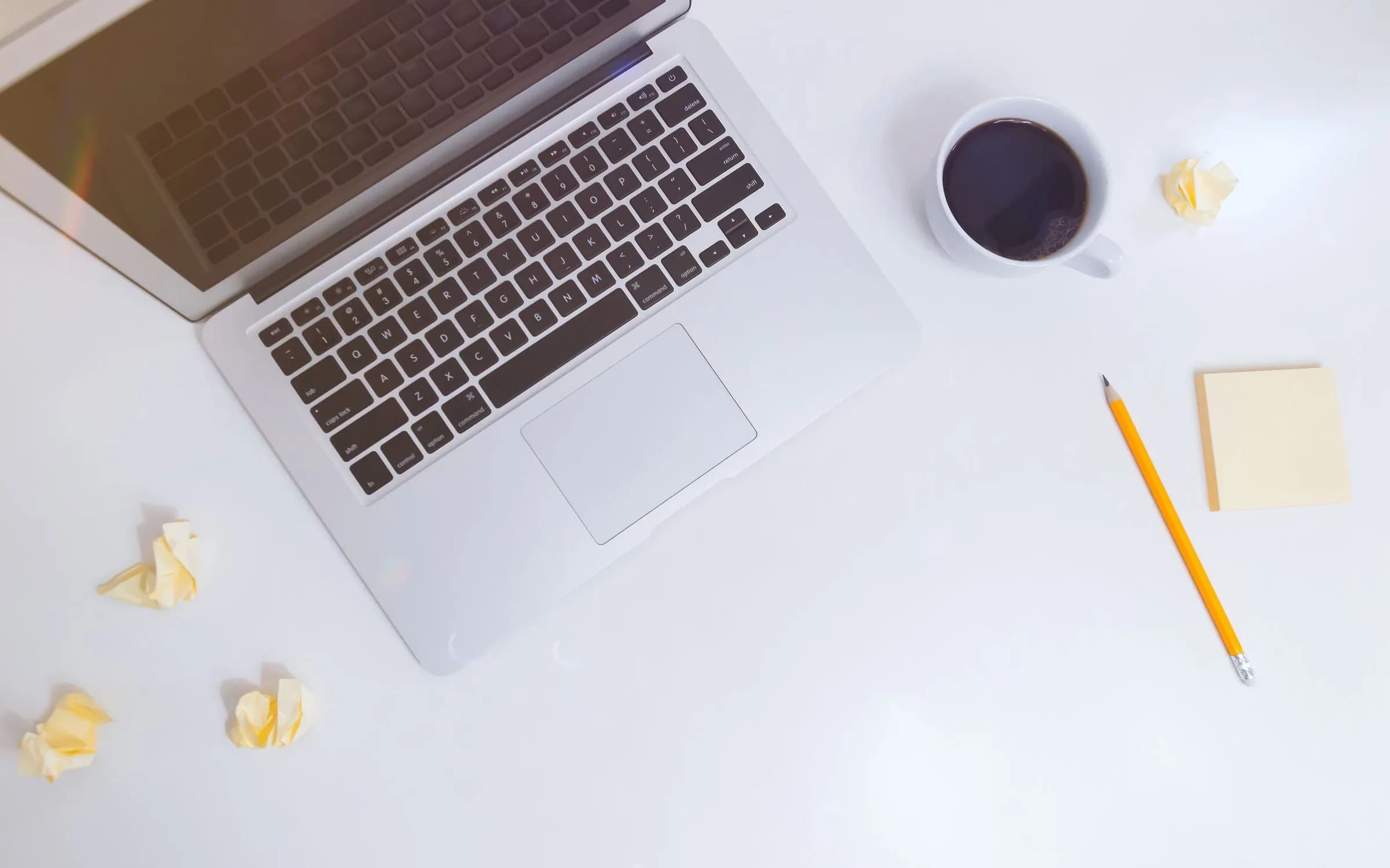 Un ordinateur portable argenté vu du dessus, posé sur un bureau blanc à côté d'une tasse de café noir, d'un crayon et d'un bloc de notes adhésives