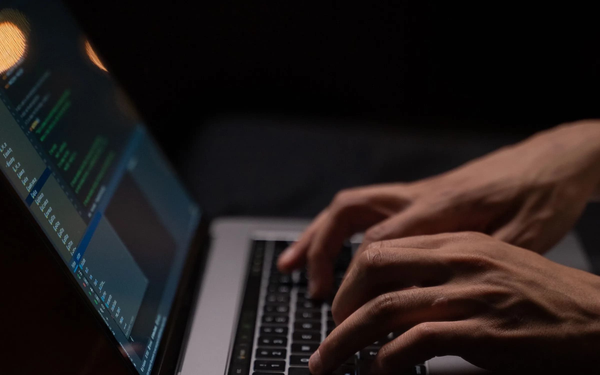 Hands typing on a laptop keyboard in a dark room, with code on the screen out of focus