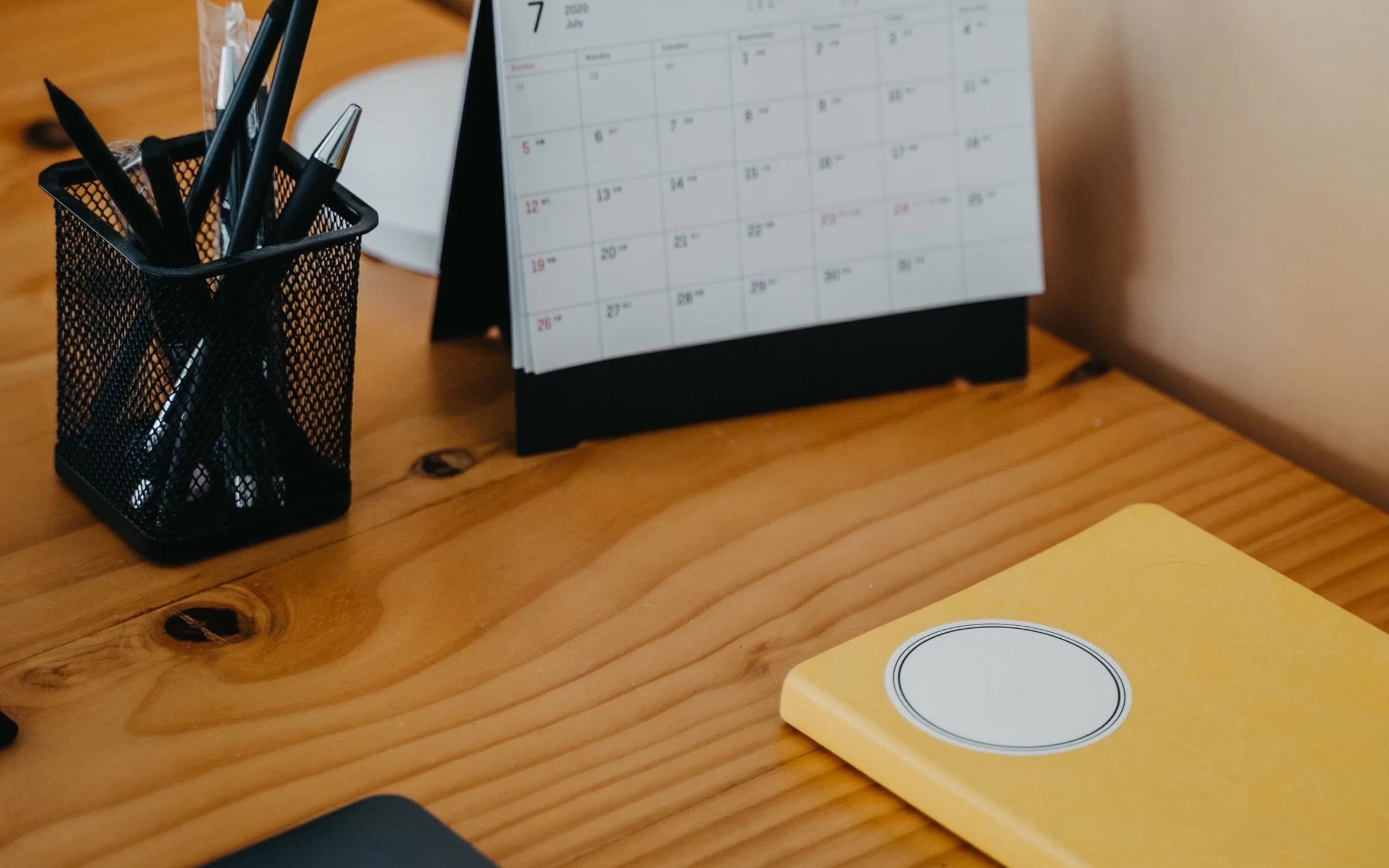 A wooden desk with a mesh pen holder, a paper desk calendar and a yellow notebook