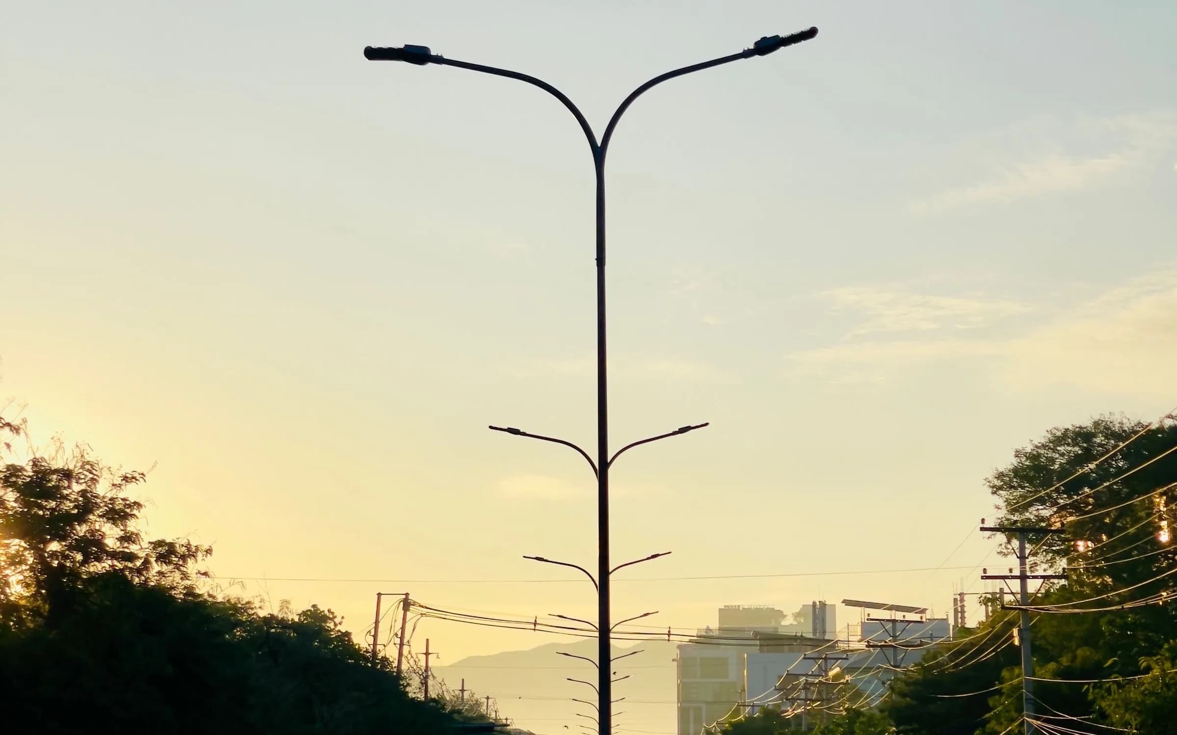 A tall double-armed street lamp silhouetted against a pale yellow sunrise sky over a quiet street
