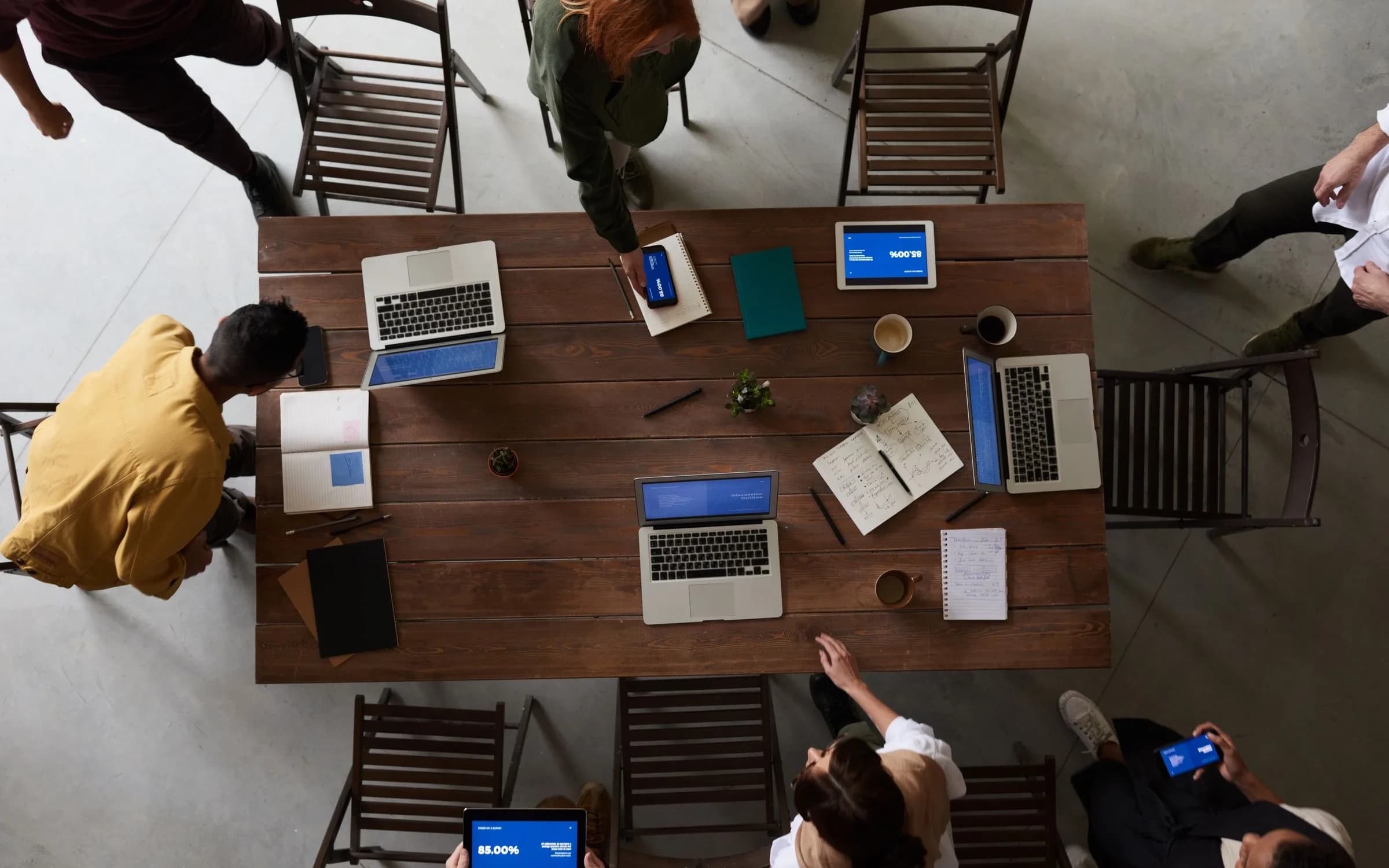 Draufsicht auf einen Besprechungstisch aus Holz mit Laptops, Notizbüchern und Kaffeetassen, an dem Kollegen gemeinsam arbeiten