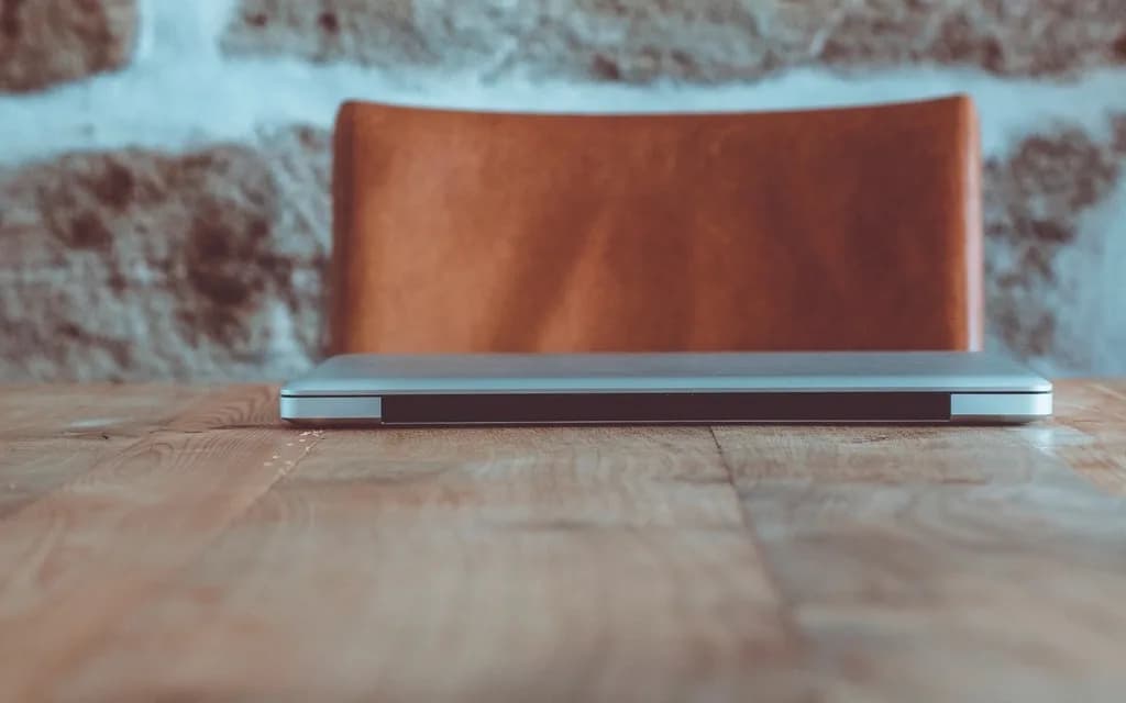 A closed silver laptop on a wooden table in front of an empty leather chair and a stone wall