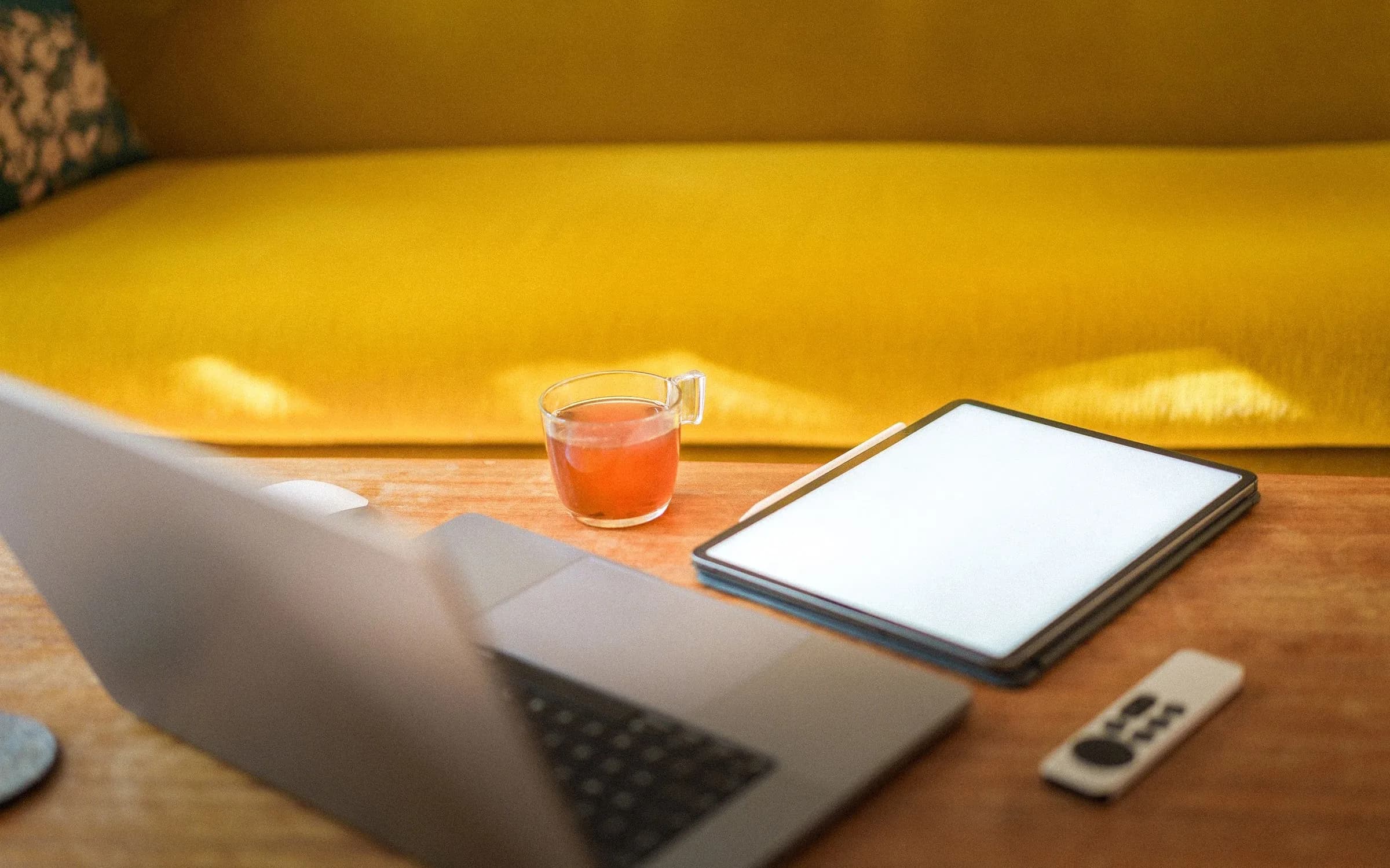 Ein Laptop, ein Tablet mit leerem Bildschirm und ein Glas Tee auf einem Holztisch vor einem gelben Sofa